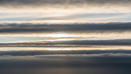 Horizontal layers of clouds are illuminated by sunlight peeking through, creating a textured sky scape with warm golden and cool blue tones.の素材