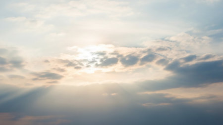Gentle sunbeams pierce through scattered clouds in a pale blue sky, creating a soft, ethereal, and hopeful atmosphere.の素材