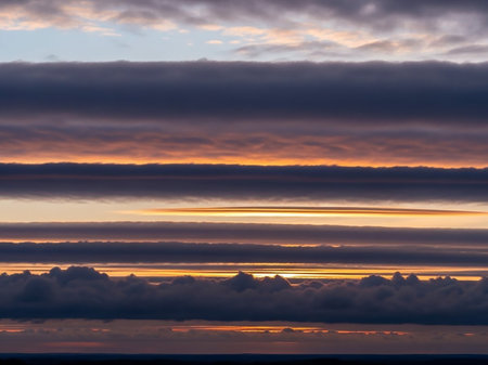 Distinct horizontal layers of dark grey and vibrant orange clouds create a striking pattern across the sky during sunset or sunrise.の素材