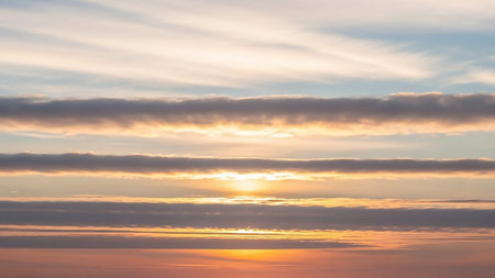 Stock showing horizontal sunset sky with multiple layers of clouds in shades of gray, orange, and yellow, illuminated by soft sunlight creating a striking and...の素材