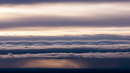 Layers of undulating gray and purple clouds stretch horizontally, with a distant golden light breaking through a rain shower, creating a dramatic, atmospheric vista.の素材