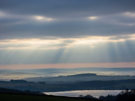 Sunbeams break through dramatic clouds over a misty landscape of rolling hills and a calm lake at dawn.の素材