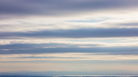 Horizontal layers of soft blue and white clouds stretch across the sky above a hazy distant landscape and a calm body of water.の素材