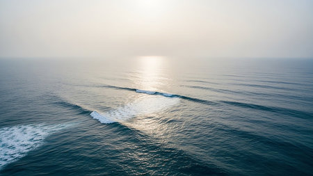Aerial view of a calm ocean with gentle waves reflecting sunlight, creating a shimmering path under a hazy, bright sky, evoking a sense of vastness.の素材
