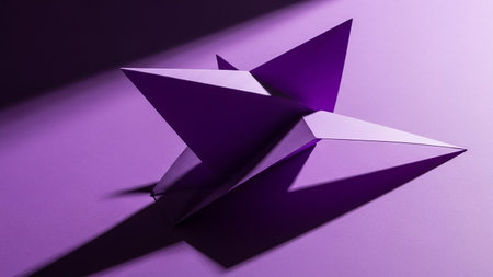 A sharp geometric origami star sculpture in deep purple casts a long, dark shadow across a textured purple surface, illuminated by dramatic studio lighting.の素材