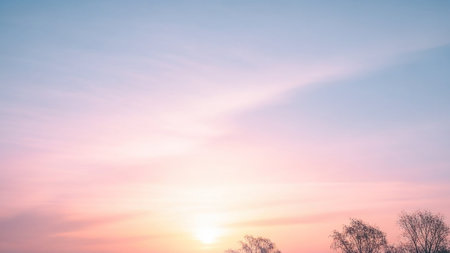 A serene sky with soft pastel pink and blue hues, featuring wispy clouds and a gentle sun glow on the horizon, with silhouetted trees at the bottom.の素材