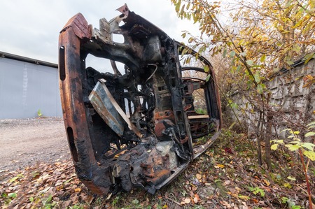 Rusty burned-out SUV on its side. Near the concrete fence on the background of trees and fallen leavesの写真素材