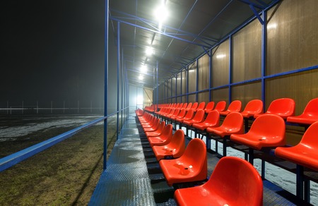Empty tribune with rows wet red plastic seats on a football field with snow and puddles. (at night)の写真素材