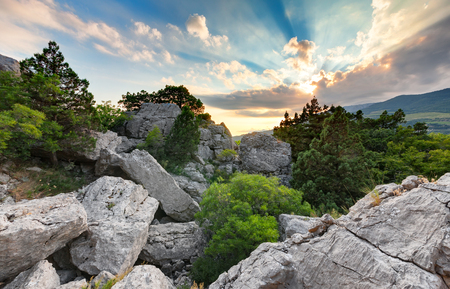 Pile of stones on the mountain among the trees and bushes in the background of the setting sun through the cloudsの写真素材