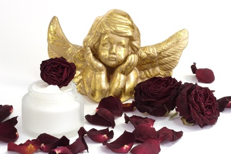 Pot of face cream, dried rose blossoms and angel figure on bright background