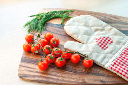 Ready to cook. Vegetables-cherry tomatoes and cooking mitten on wooden cooking board. Indoors.の写真素材