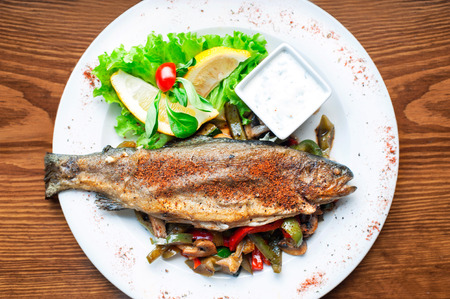 Grilled trout with fried vegetables salad and lemon on white porcelain plate. Main course. Indoors.の写真素材