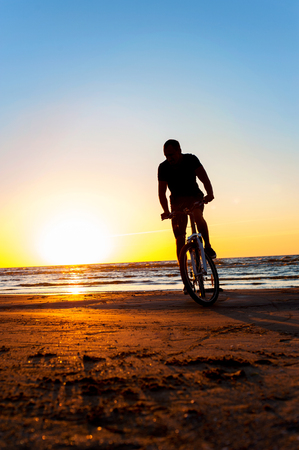 Young man cyclist silhouette on blue sky and sunset background on the beach. Summertime multicolored outdoors vertical image.の写真素材