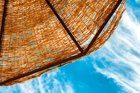 Wattled straw beach umbrella on blue cloudy sky background. Outdoors summertime multi colored closeup horizontal image. View from below.の写真素材