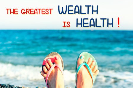 The greatest wealth is health. Motivational inspirational quote with funny cute tidy woman's feet on the beach on ocean waves background. Colored horizontal summertime image with vintage filter.の写真素材
