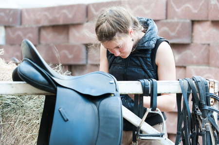 Pretty teenage girl equestrian cleans black Leather Horse Saddle and equipment at farm. Horizontal outdoors summertime image.の写真素材
