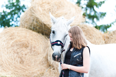 Portrait of pretty young blondy teenage cheerful girl owner with her favorite white horse at farm yard on yellow hay/straw rolled stack background. Vibrant colored outdoors horizontal summertime image.の写真素材