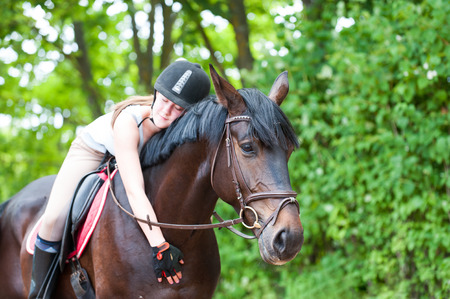 Young cheerful teenage girl-equestrian with closed eyes tenderly embracing her big lovely brown horse. Vibrant multicolored summertime outdoors horizontal image.の写真素材
