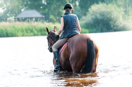 Young teenage lady equestrian riding horseback in river at early morning in rays of sunlight. View from backside. Multicolored vibrant outdoors horizontal summertime image.の写真素材