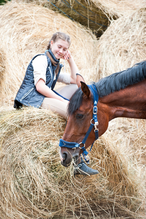 Pretty young teenage girl owner sitting on yellow hay/straw rolled stack and looking after her favorite bay horse at farm yard. Vibrant colored outdoors vertical summertime image.の写真素材