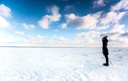 One girl standing at the edge of frozen sea and looking away contemplating the frozen waves in winter time. Outdoors horizontal image with cloudy blue sky background. Wide angle panoramic viewの写真素材