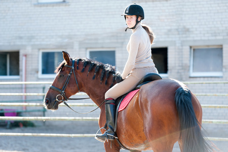Young teenage girl riding a horseback at equestrian farm after sport training. Colored outdoors horizontal summertime image. View from backside.の写真素材