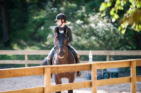 Young pretty cheerful girl resting after horseback riding training on arena. Vibrant colored...