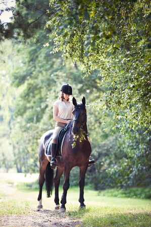 Young teenage girl equestrian riding horseback at spectacular early morning in rays of sunlight. Green trees frame. Multicolored vibrant outdoors vertical summertime image with vintage filterの写真素材