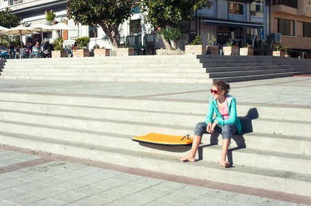 Mediterranean lifestyle. Girl with surfboard sitting on city main square Tenerife, Canary islands, Spain, El Medano. Multicolored summertime outdoors horizontal image.の写真素材