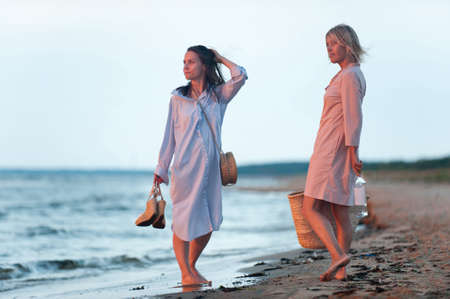 Two young women walking along the seacoast at sunset. Friends/sisters. Summertime outdoors inspirational horizontal image with vintage filter. Spectacular cloudy sky backgroundの写真素材