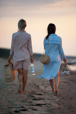 Two young women walking along the seacoast at sunset. Friends/sisters. Summertime outdoors inspirational vertical image with vintage filter. Spectacular cloudy sky background. View from backsideの写真素材