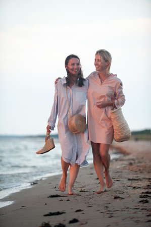 Two young women walking along the seacoast at sunset. Friends/sisters. Summertime outdoors inspirational vertical image with vintage filter. Spectacular cloudy sky backgroundの写真素材