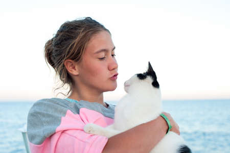 Portrait of tanned teenage girl kissing cat on pier. Colored outdoor horizontal image with blue seascape  background.の写真素材