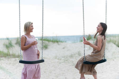 Two young attractive women resting and drinking white wine on the beach. Friends/sisters. Summertime outdoors inspirational horizontal image.の写真素材