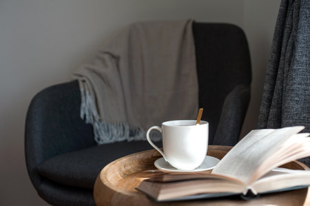 Cozy textile armchair with warm blanket and opened book with ceramic cup of tea on wooden coffee table for relaxing evening. Lifestyle indoor horizontal colored image.の写真素材