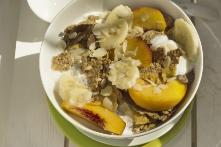Breakfast in the sun, cereal bowl, peach and banana, yogurt on green plate and warm orange color. White on woodの写真素材