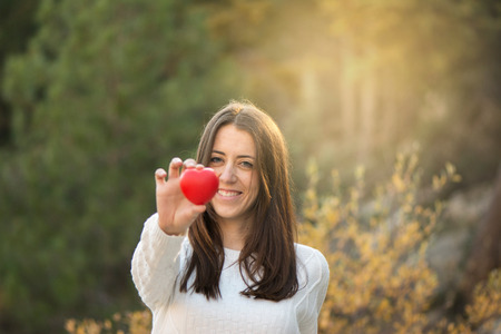 Portrait of beautiful young woman holding red heart against of forestの写真素材