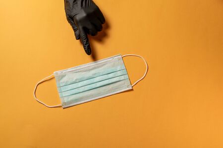 Hand in black protective glove, points out Typical 3-ply surgical mask to cover the mouth and nose with text space on orange background, Virus prevention and quarantine conceptの写真素材