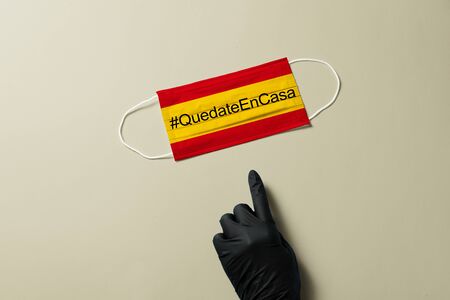 COVID-19, #QuedateEnCasa. Hand in black protective glove, points out surgical mask with the Spain flag showing the message "#QUEDATE EN CASA"., Virus prevention and Spain quarantine conceptの写真素材