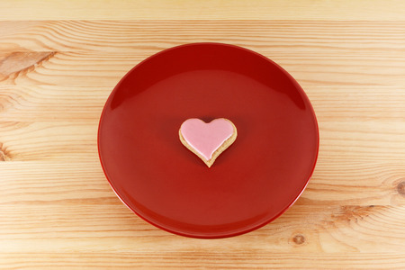 One heart-shaped cookie with pink frosting on a red plate on a wooden tableの写真素材
