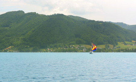 Sailing boat with a colourful sail on Lake Attersee in the Salzkammergut, Austria - clear blue water and green wooded hillsのeditorial素材