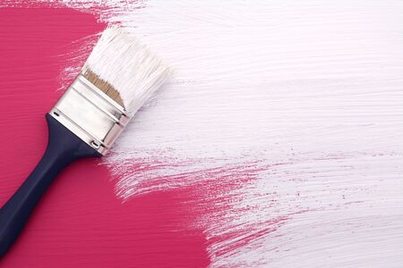 Paintbrush with white paint painting over deep pink colour on a pine boardの写真素材