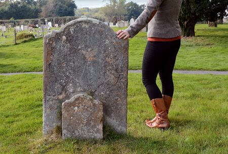 Smartly dressed woman rests her hand thoughtfully on a gravestone in a cemeteryの写真素材