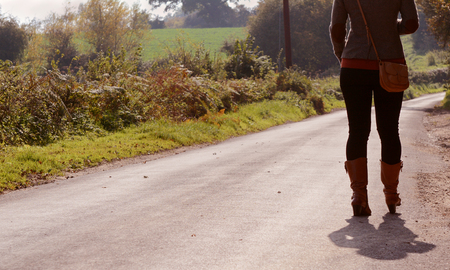 Fashionably dressed young woman walks up a country lane alone in autumn, heading into the sunの写真素材