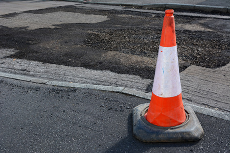 Bright orange and white traffic cone on a pavement, damaged road surface beyond, undergoing repair...