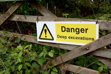 Warning sign - Danger Deep Excavations - fixed to a rustic wooden gate, overgrown with nettles in...