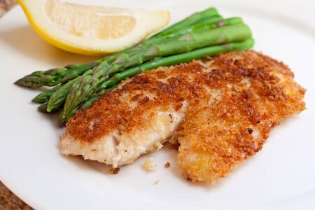 Closeup of fried breaded tilapia fillet with asparagus and lemon on white plate