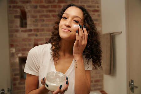 Multi-cultural young adult female happily applying moisturiser on her face in her bathroom mirrorの写真素材