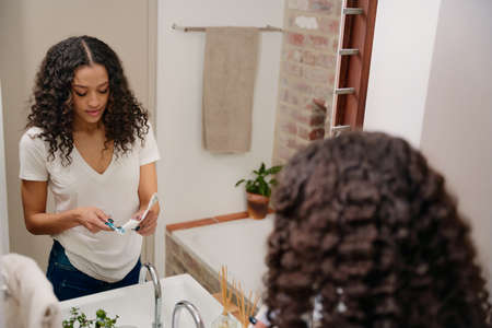 Mirror Reflection of a Young Beautiful African American Woman Putting Toothpaste on her ToothBrush in the Bathroomの写真素材