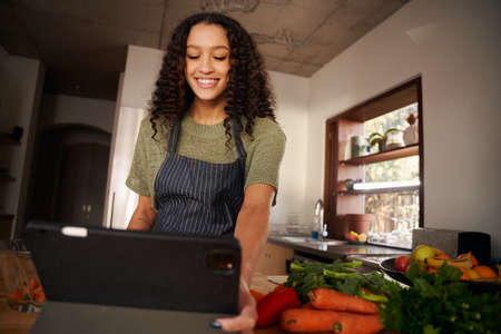 Low angle of Multi-cultural young female reading recipe from tablet while cooking in modern kitchen....